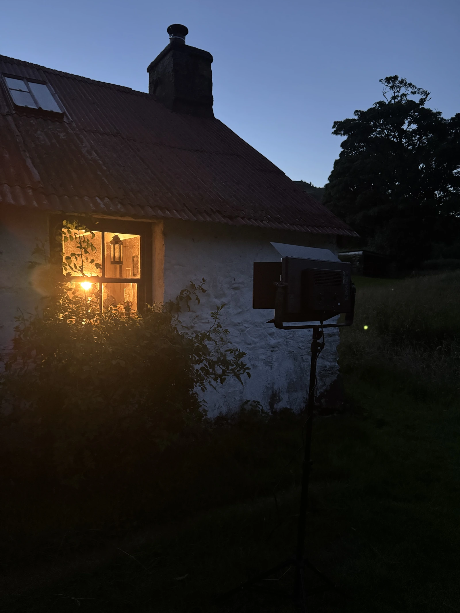 Nächtlicher Drehaufbau: Filmleuchte auf einem Stativ vor einem Cottage, warmes Licht im Fenster